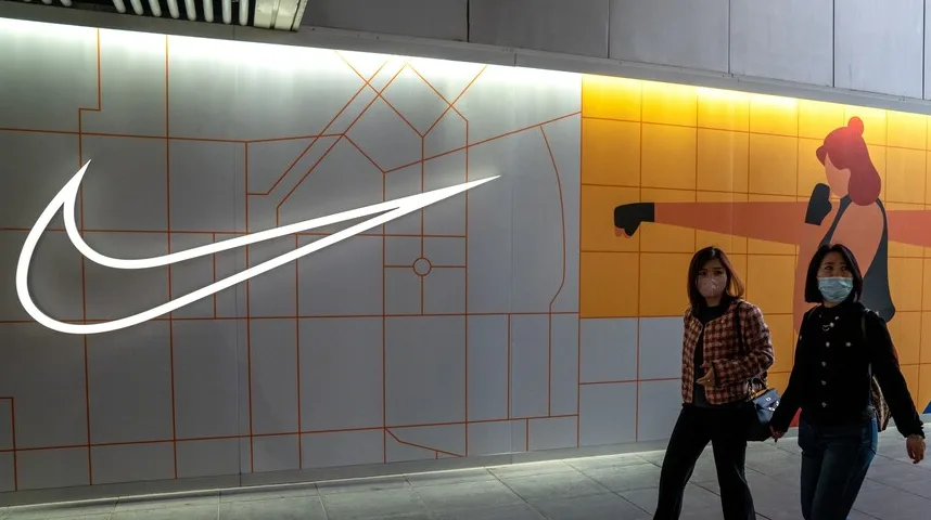 Dva ženske osobe nose maske za lice dok prolaze pored velikog osvijetljenog Nike znaka.