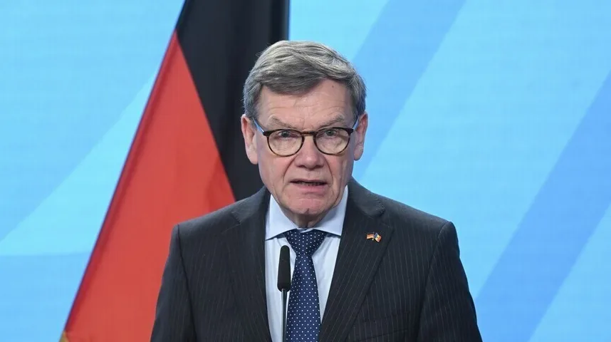 Portret starijeg muškarca sa naočarima kako govori u mikrofon, sa njemačkom zastavom u pozadini.