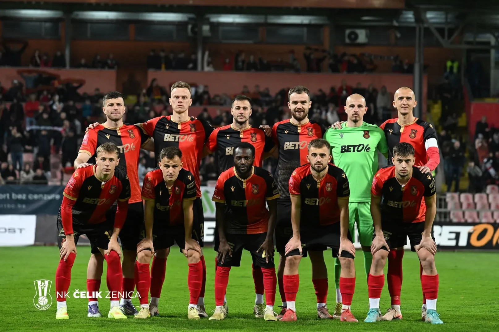 Fudbalski tim NK Čelik Zenica pozira za timsku fotografiju na terenu, obučeni u crno-crvene dresove.