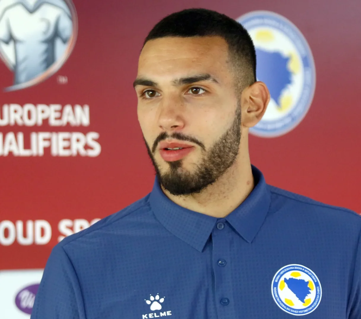 Portret mladog nogometaša u plavom dresu sa logom Nogometnog/Fudbalskog saveza Bosne i Hercegovine i natpisom "European Qualifiers" u pozadini.