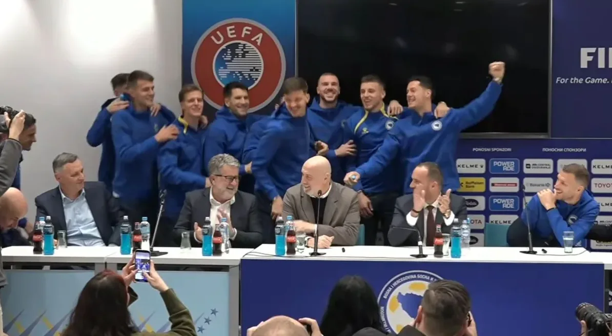 Nogometna reprezentacija Bosne i Hercegovine slavi uzdignutih ruku u dvorani s logom UEFA-e i FIFA-e u pozadini.