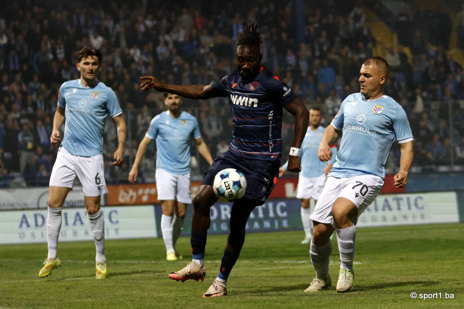 Fudbaler u tamnoplavom dresu izvodi dribling sa loptom, okružen igračima u plavim dresovima.