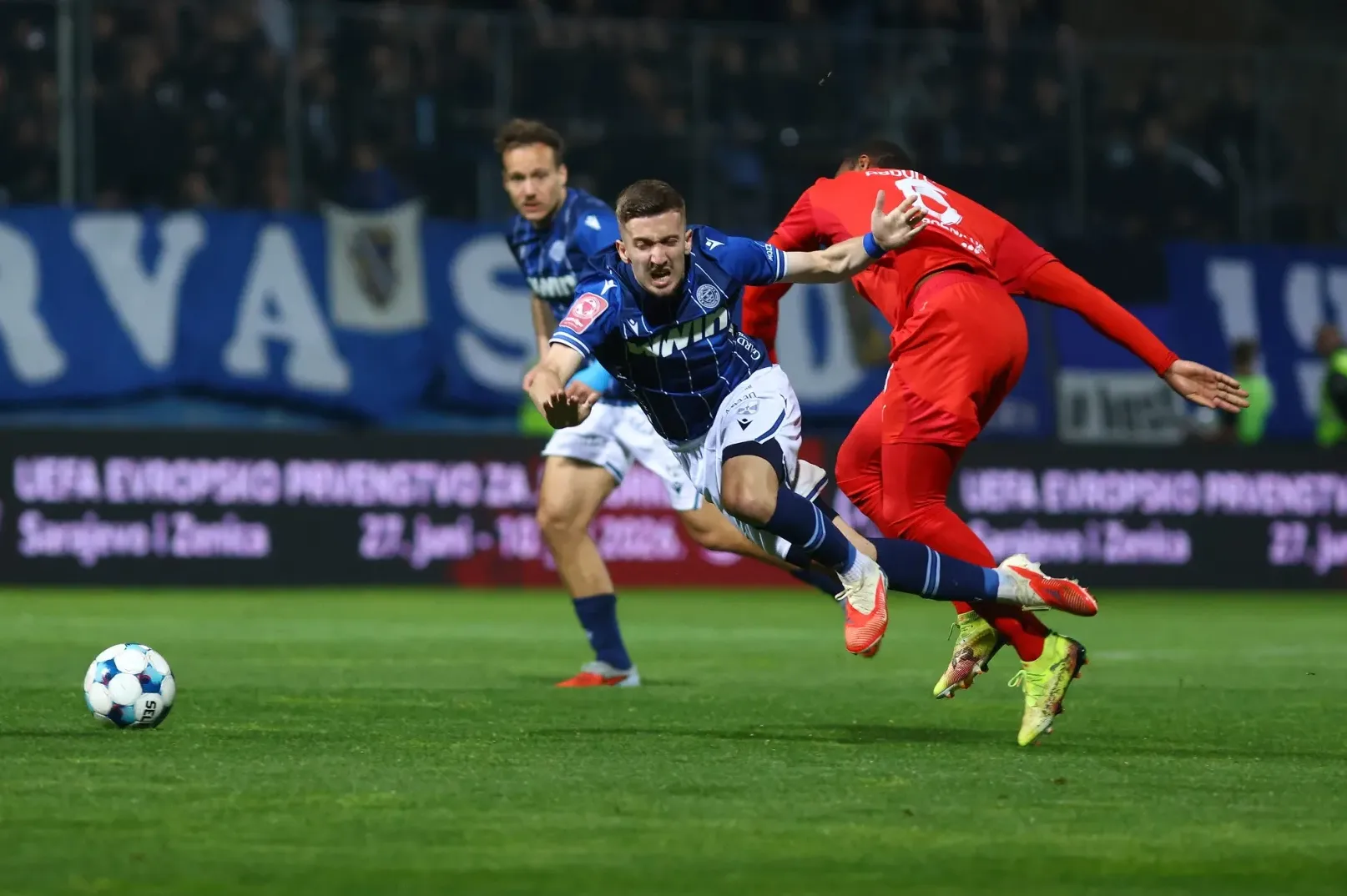 Fudbaleri u plavim i crvenim dresovima u borbi za loptu na zelenom terenu.