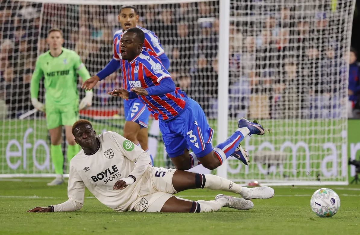 Fudbaler u bijelom dresu pada na travnjak, dok ga fudbaler u plavo-crvenom dresu dodiruje nogom.