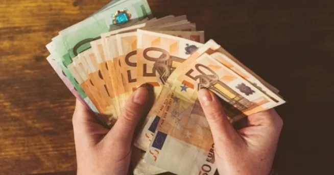 Ruke drže snop novčanica eura, pretežno apoena od 50 eura, sa primjetnom zelenom novčanicom u pozadini.