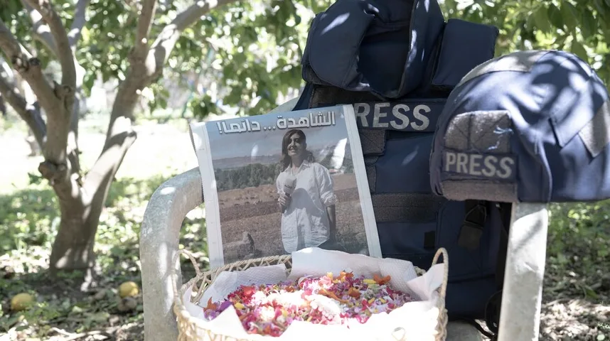 Prsluk i kaciga sa natpisom 'PRESS' pored novina sa slikom novinarke i korpe sa cvijećem.