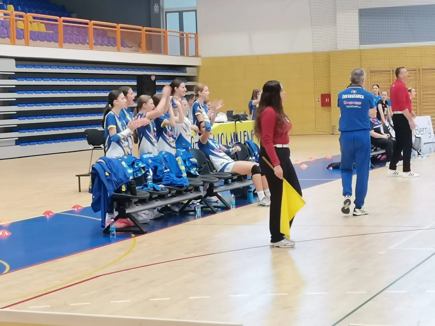 Sportistkinje u plavim dresovima sjede na klupi i aplaudiraju, dok sudije stoje na terenu sa žutim zastavicom.