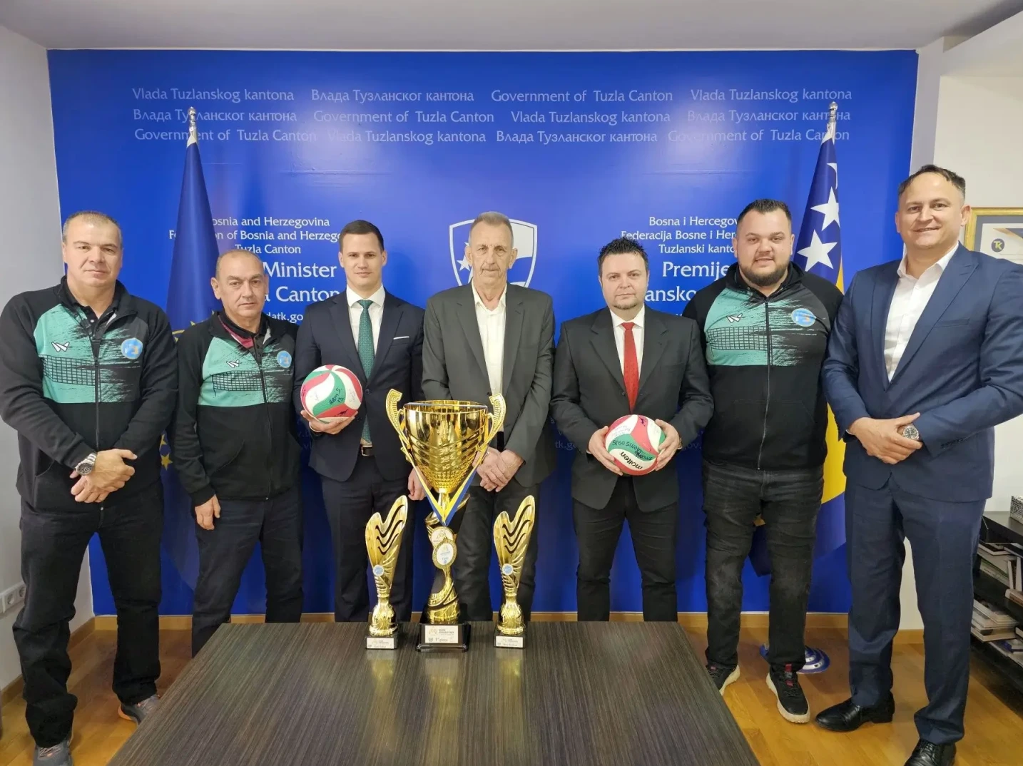 Grupa muškaraca u odijelima i sportskim trenerkama pozira sa odbojkaškim loptama i peharima ispred plave pozadine sa logotipima Vlade Tuzlanskog kantona.