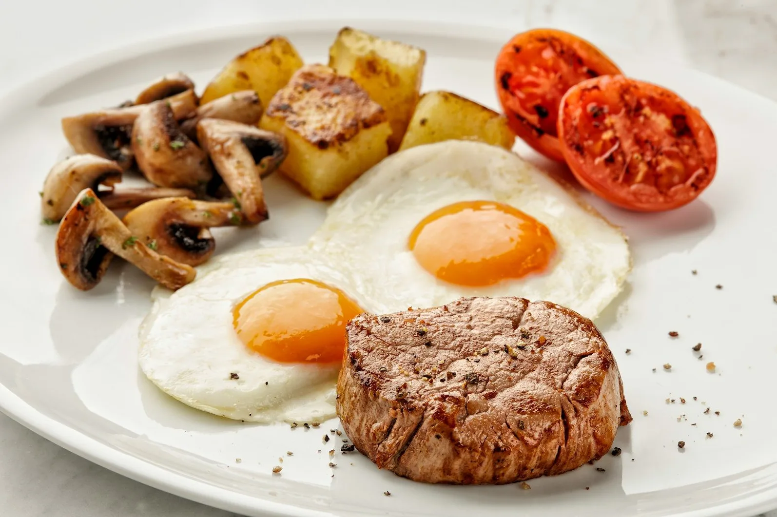 Odrezak, dva jajeta na oko, pečene gljive, kriške pečenog krompira i polovine paradajza servirani na bijelom tanjiru.