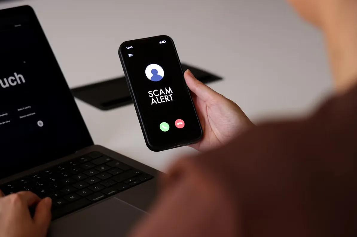 Ruka drži pametni telefon na ekranu kojeg piše 'Scam Alert' (Upozorenje o prevari) i prikazuje dugmad za javljanje i odbijanje poziva, pored laptopa.