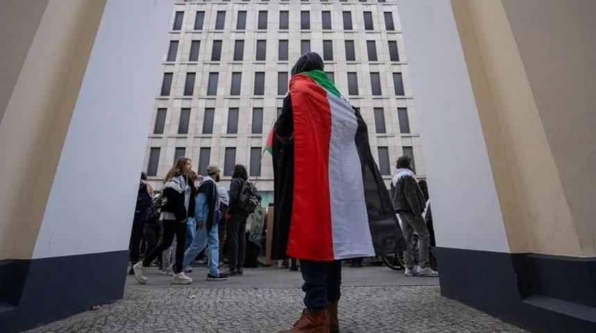 Osoba ogrnuta palestinskom zastavom stoji okrenuta leđima kameri, dok se iza nje vidi grupa ljudi i moderna zgrada.