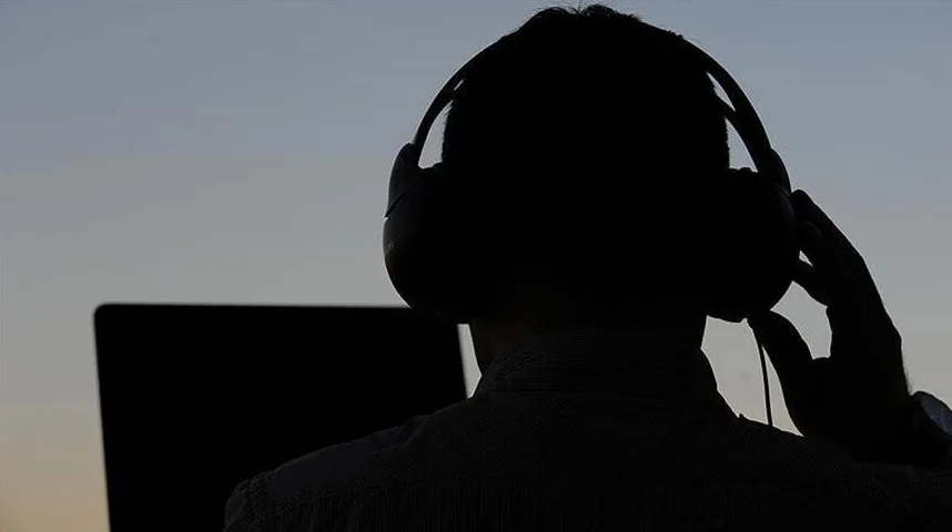 Silueta osobe sa slušalicama na glavi i rukom na uhu, ispred tamnog ekrana laptopa, sa nejasnom pozadinom zalaska sunca.