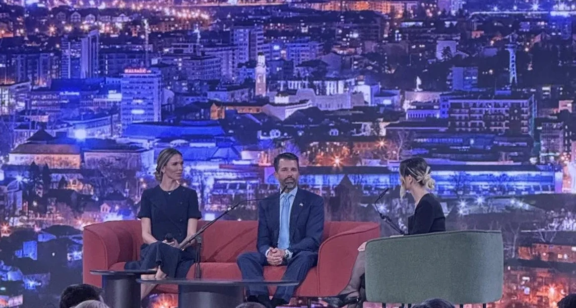 Tri osobe sjede na crvenoj sofi i zelenoj fotelji ispred pozadine s osvijetljenim gradskim pejzažom noću.