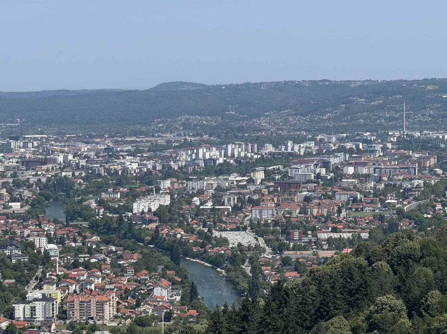 Panoramski pogled na gradsku panoramu sa urbanim zgradama, rijekom koja protiče kroz grad i brdima u pozadini.