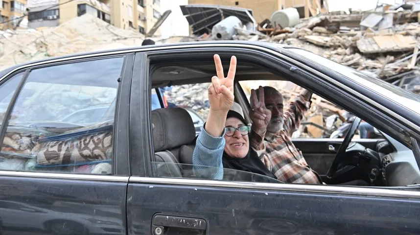Žena i muškarac u automobilu podižu dva prsta u znak pobjede, dok se u pozadini vide ruševine zgrada.