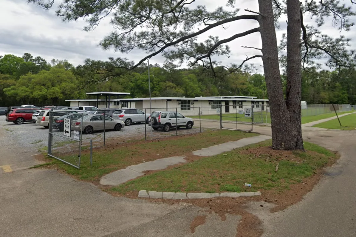 Parking lot pun autoškolskih vozila ispred niske školske zgrade s metalnom ogradom i drvećem.