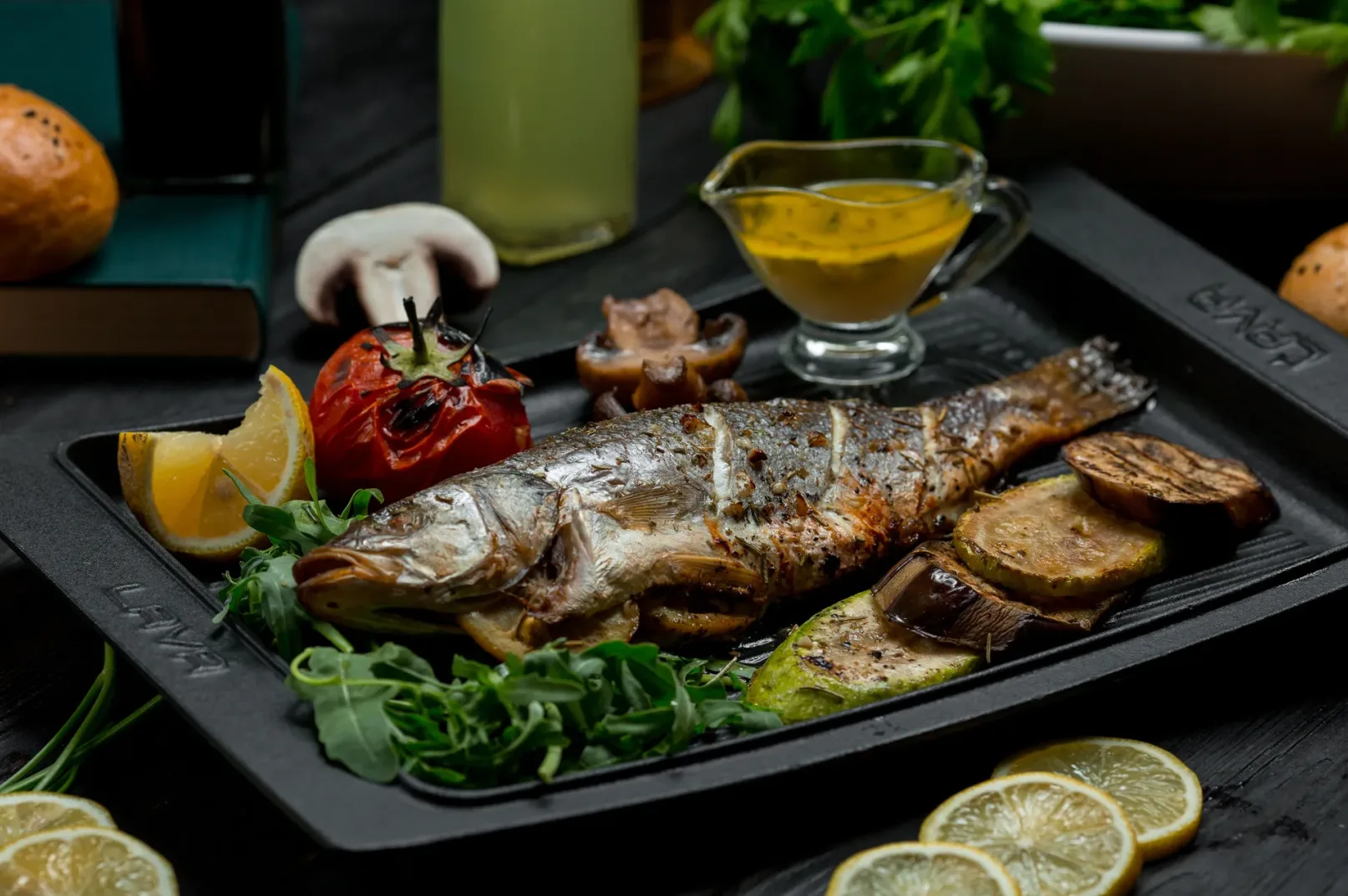 Pečena cijela riba servirana na crnom tanjiru sa grilovanim povrćem (tikvice, patlidžan), kriškom limuna, grilovanim paradajzom i listovima rukole. U pozadini se nalazi činija sa sosom i piće.