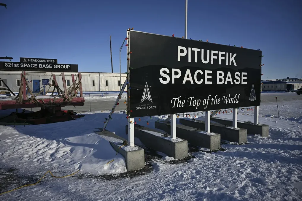 Natpis 'PITUFFIK SPACE BASE' iznad natpisa 'The Top of the World' sa logom američkih svemirskih snaga, na snijegom pokrivenom terenu sa zgradama u pozadini.