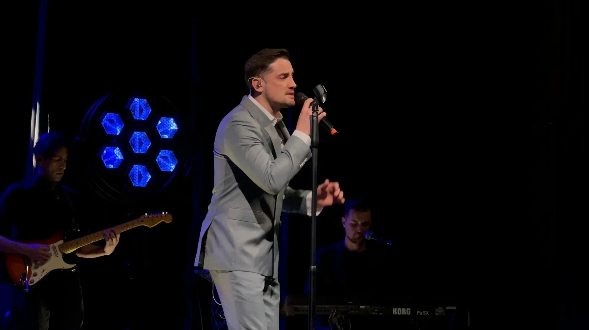 Mladić u svijetlosivom odijelu pjeva na bini uz pratnju benda.
