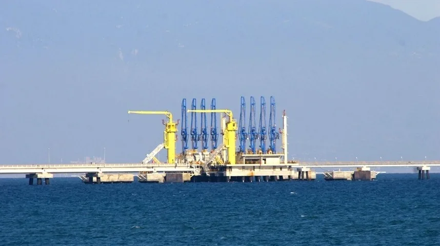 Industrijska platforma na moru sa nizom plavih dizalica i žutim podupiračima, povezana sa kopnom dugim mostom.