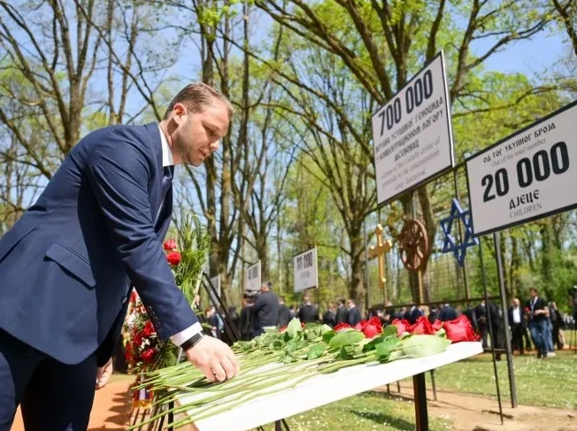 Čovjek u tamnom odijelu polaže crvene ruže na sto prepun cvijeća ispred spomen-obilježja žrtvama u Jasenovcu.