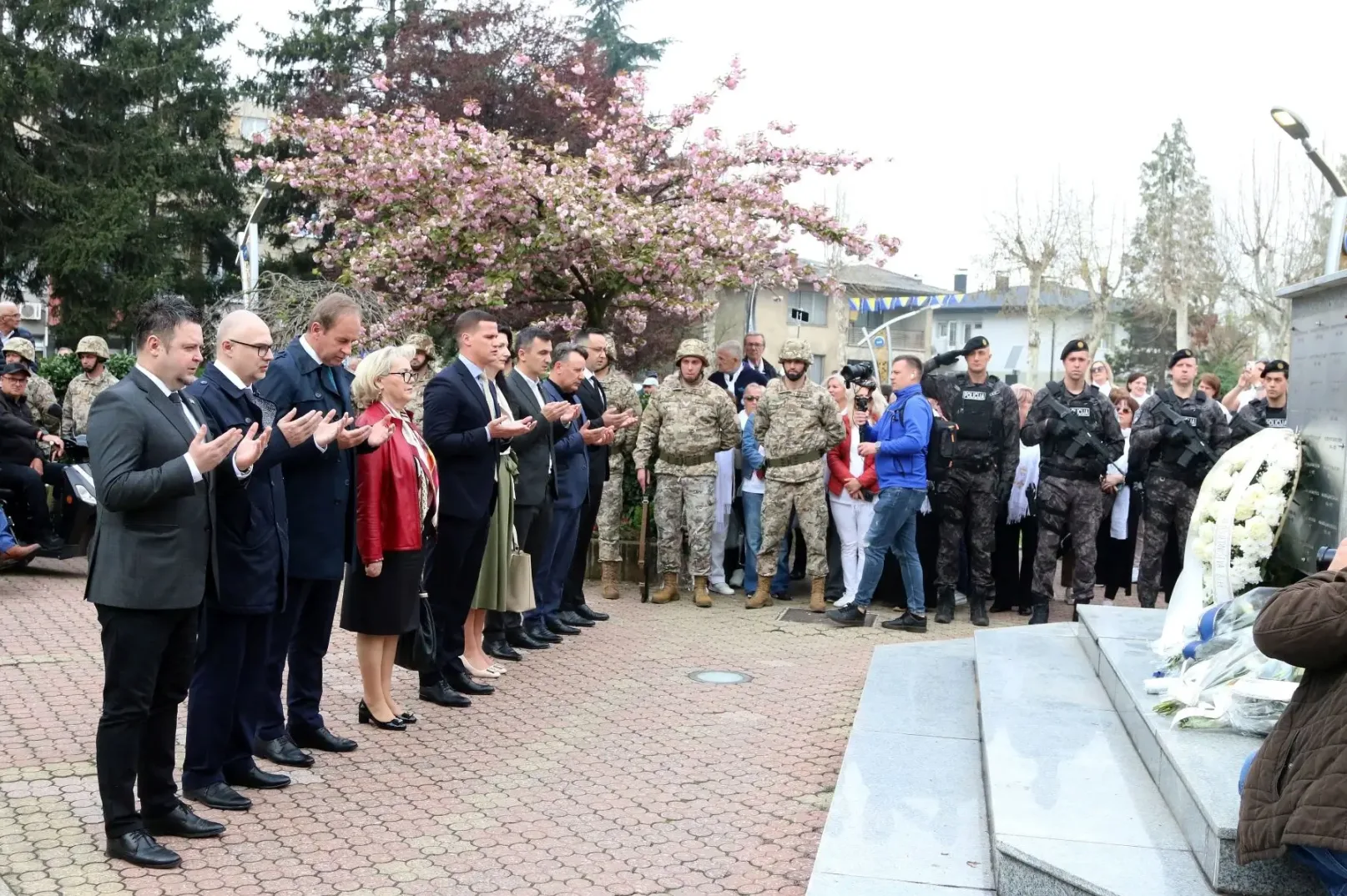 Ljudi u odijelima i uniformama polažu cvijeće na spomen-obilježje u blizini drveta s ružičastim cvjetovima.