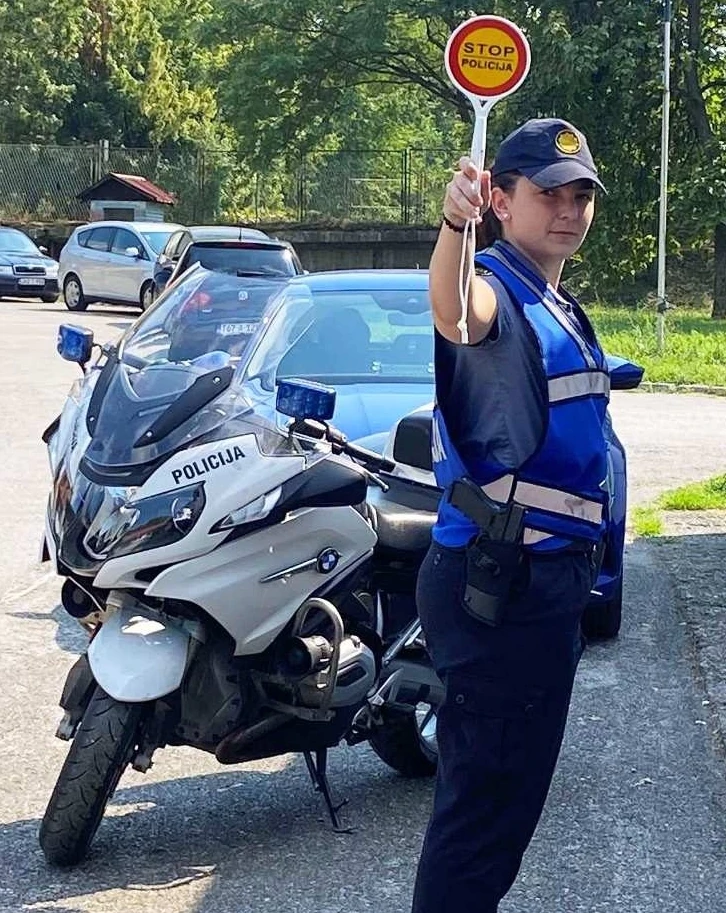 Policajka saobraćajne policije drži znak "STOP POLICIJA" stojeći pored policijskog motocikla.