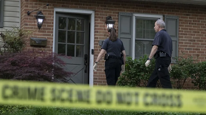 Dva policijska službenika, jedan muškarac i jedna žena, pregledavaju ulaz u kuću dok je žuta traka "CRIME SCENE DO NOT CROSS" postavljena ispred.