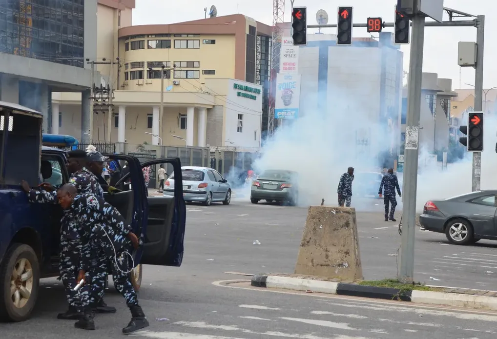 Policijski službenici u uniformama stoje pored policijskog vozila dok se dim suzavca širi ulicom.