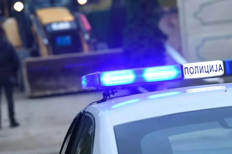 Plava rotacija na krovu policijskog vozila sa natpisom "ПОЛИЦИЈА" u pozadini sa bagerom i osobom.