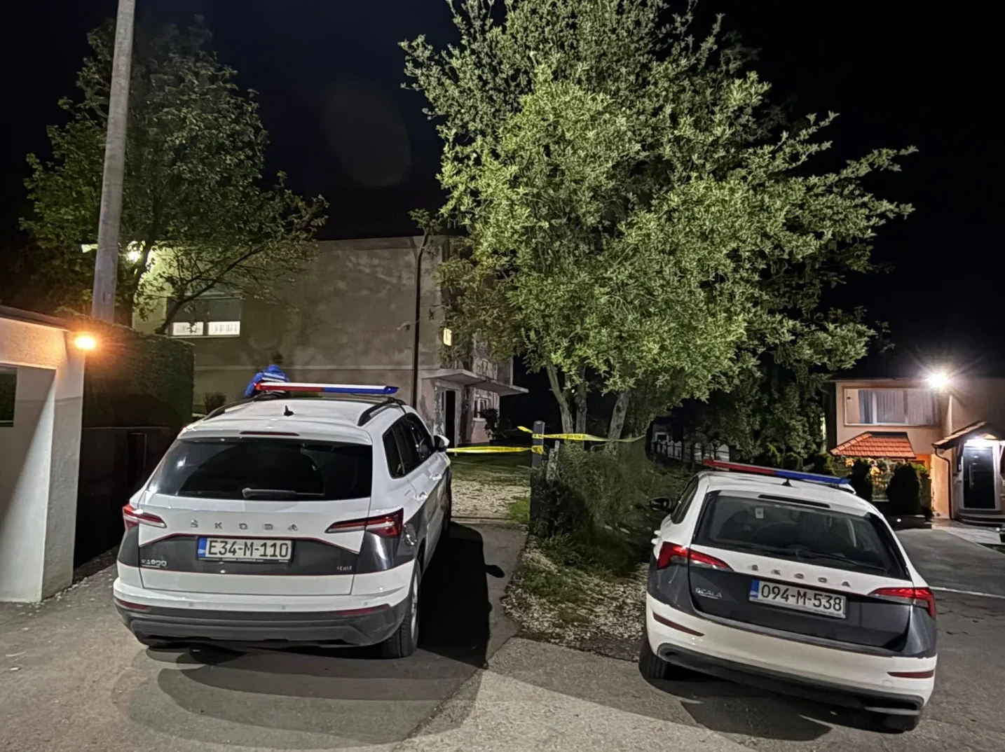 Dva policijska automobila parkirana ispred kuće noću, sa žutom policijskom trakom koja ograđuje prostor.