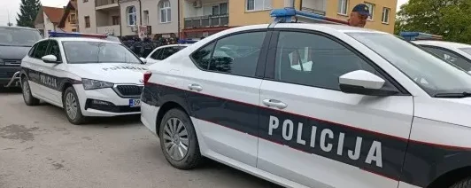 Bijela policijska vozila s natpisom "POLICIJA" parkirana na ulici, s policajcem koji viri iznad jednog od vozila.