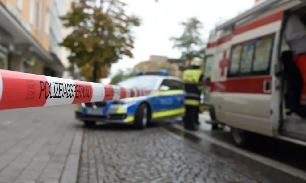 Policijska traka sa natpisom "POLIZEIABSperrung" u prvom planu, policijsko vozilo sa upaljenim rotacijama i vozilo hitne pomoći u pozadini.