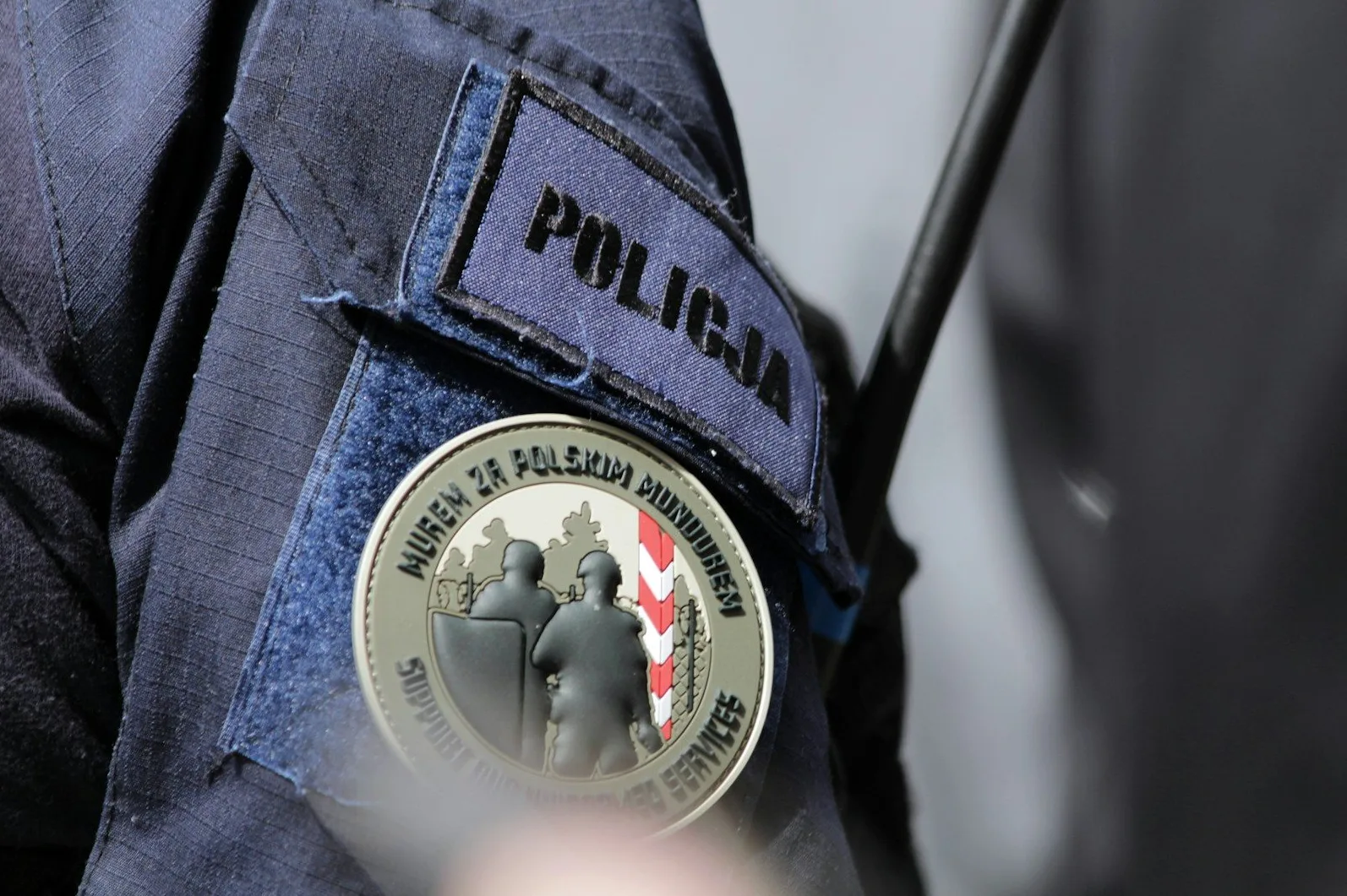 Krupni kadar na policijsku uniformu sa grbom "Murem za Polskim Mundurem" koji prikazuje dva policajca u zaštitnoj opremi i poljske nacionalne boje.