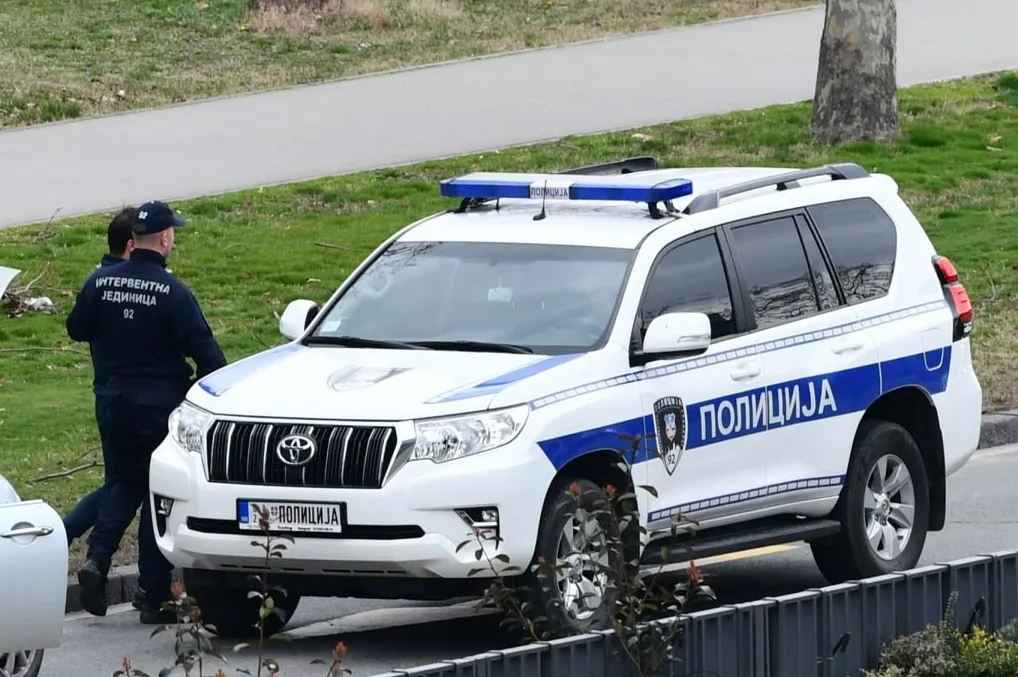 Bijelo policijsko vozilo sa plavim svjetlosnim signalima i oznakama "POLICIJA", pored kojeg stoje službenici u tamnoj uniformi.