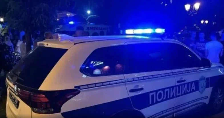 Policijsko vozilo sa upaljenim plavim rotirkama, u pozadini se vide ljudi i zgrada hitne pomoći.