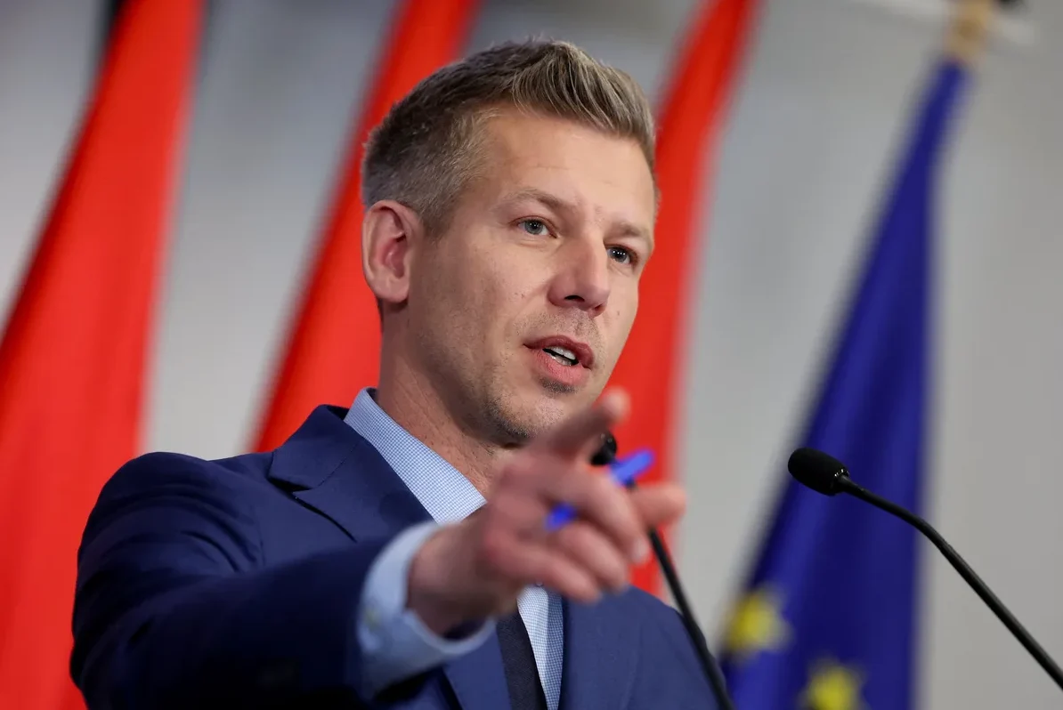 Političar u plavom odijelu pokazuje prstom ispred mikrofona, sa zastavama iza njega.