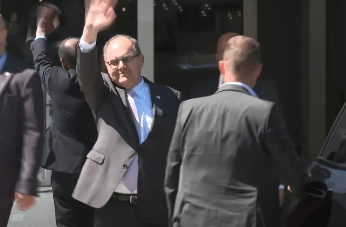 Političar u tamnom odijelu i plavoj košulji sa ljubičastom kravatom podignutih ruku maše dok izlazi iz zgrade u pratnji osiguranja.