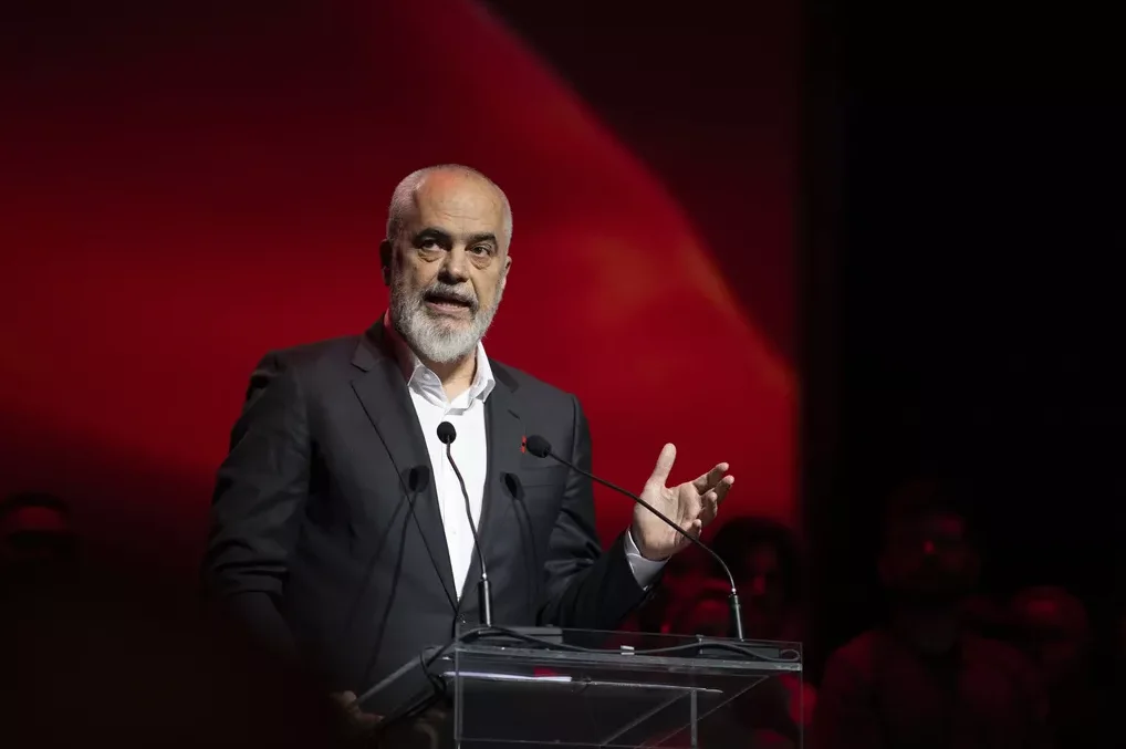 Premijer Albanije Edi Rama govori sa govornice, držeći ruke uzdignute dok govori pred publikom osvijetljenom crvenom pozadinom.