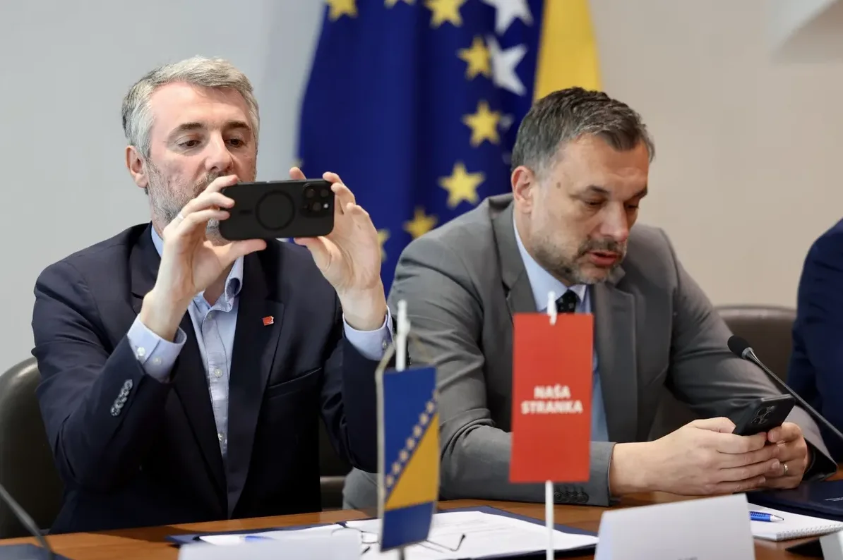 Političar u plavom sakou drži pametni telefon u horizontalnom položaju i snima nešto ispred sebe, dok drugi političar u sivom odijelu gleda u svoj telefon.