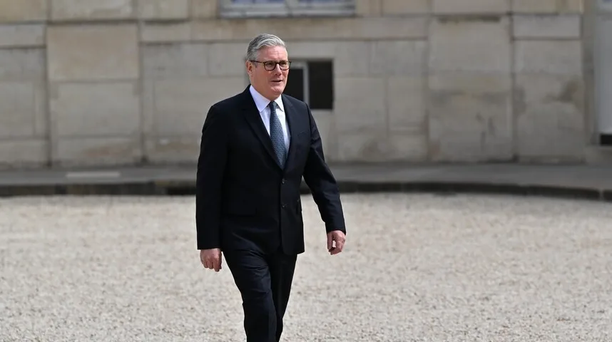 Keir Starmer u tamnom odijelu, bijeloj košulji i plavoj kravati, nosi naočale, hoda po šljunku ispred zgrade.