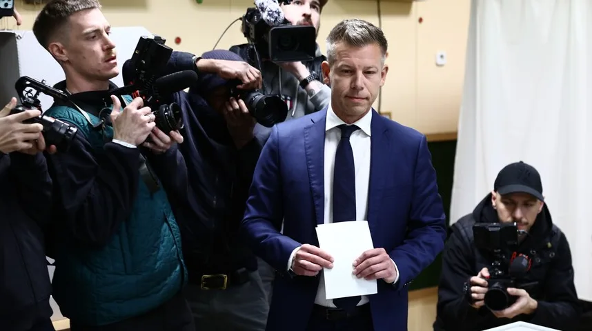 Političar u tamnoplavom odijelu i tamnoplavoj kravati drži bijeli papir u ruci, dok ga okružuju novinari s kamerama i fotoaparatima.