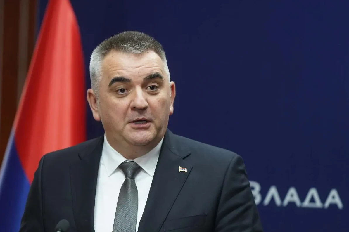 Političar u tamnom odijelu i bijeloj košulji sa sivom kravatom, koji govori ispred zastave sa crvenom i plavom bojom, te natpisom "ВЛАДА" na tamnoplavoj pozadini.