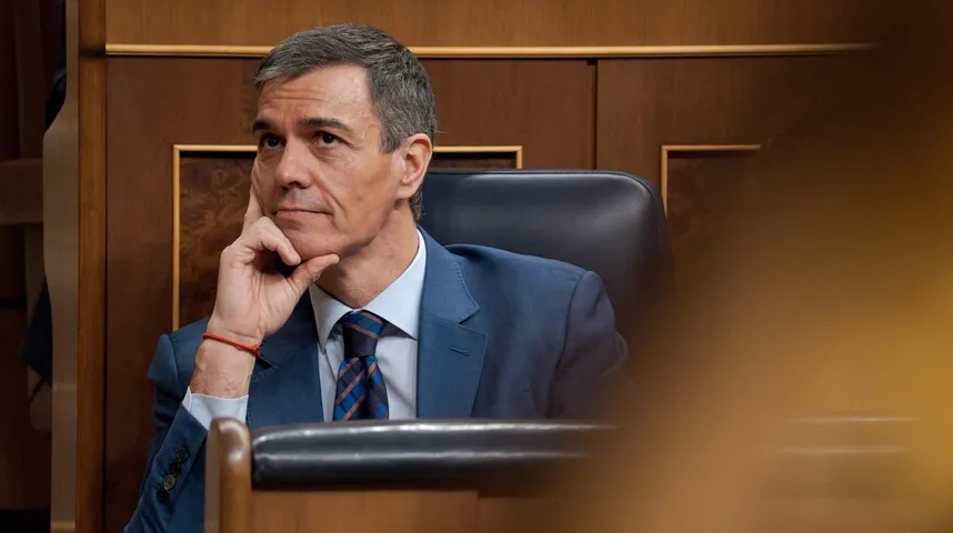 Političar u plavom odijelu i plavo-smeđoj kravati sjedi u stolici, oslanjajući bradu na ruku.