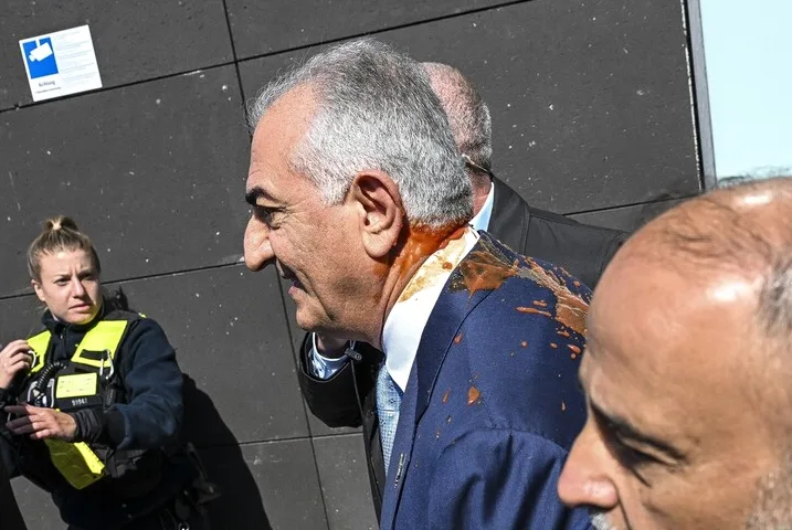 Muškarac u tamnom odijelu i plavoj košulji s plavom kravatom, sa vidljivim sivim vlasima, stoji okrenut profilom prema lijevoj strani slike. Njegov kaput i vrat su obilno isprskani crvenkastom tečnošću, vjerovatno kečapom ili nekim drugim sosom. Iza njega se vide obrisi još dvojice muškaraca, od kojih je jedan takođe u odijelu. Na lijevoj strani slike, ispred njega, stoji žena u tamnoj uniformi sa žutim prslukom visoke vidljivosti, pokazujući zabrinutost na licu.