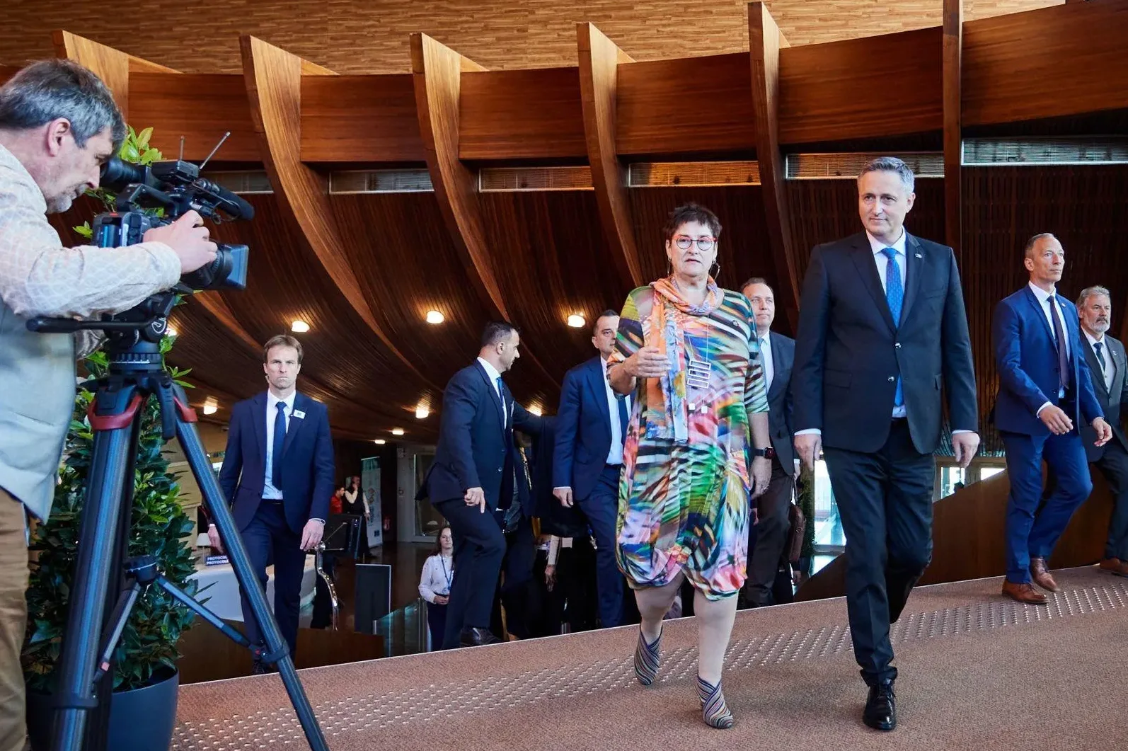 Političari u tamnim odijelima i žena u šarenoj haljini koračaju prema kameri, dok ih snimatelj snima