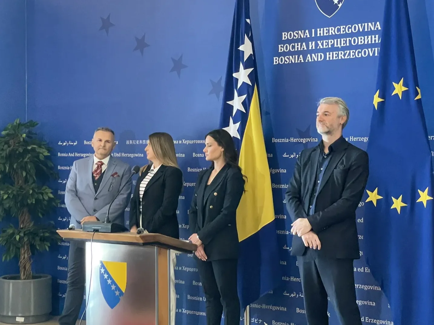 Četiri osobe stoje ispred zastave Bosne i Hercegovine i Evropske unije. Jedna osoba stoji za podijumom sa grbom BiH.