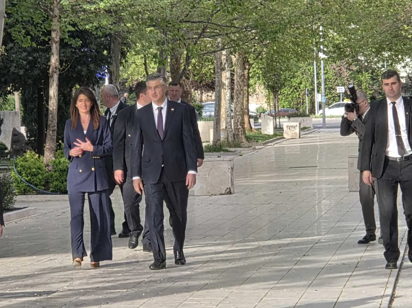 Premijer Andrej Plenković i ministrica vanjskih poslova Republike Hrvatske s pratnjom hodaju kamenim pločnikom u vanjskom prostoru uz drvored.