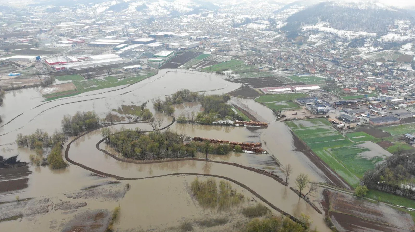 Aerialni snimak poplavljenog naseljenog mjesta, industrijskih objekata i poljoprivrednog zemljišta sa brdima prekrivenim snijegom u pozadini. Rijeka je izašla iz korita i preplavila gradske ulice i okolne njive.