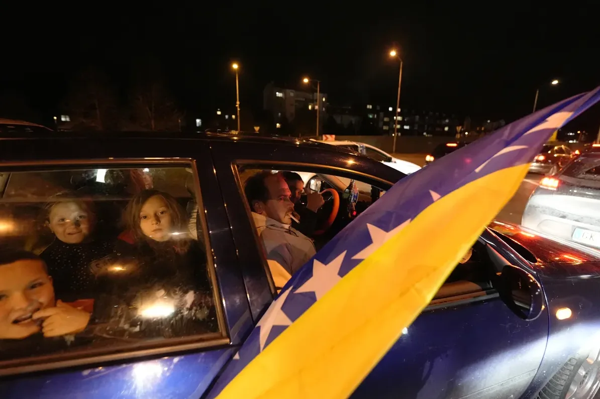 Djeca gledaju kroz prozor automobila dok se voze kroz grad noću, sa zastavom Bosne i Hercegovine istaknutom u prvom planu.